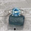 The "Arethusa" ring features a 2.65-carat Montana sapphire set in platinum, shown on a textured blue mineral background with the "Earth's Treasury" logo below.