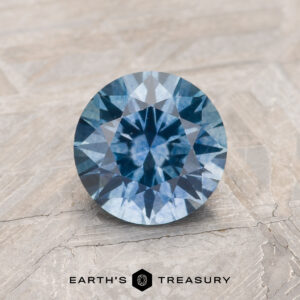 A 1.33-carat medium blue heated Montana Sapphire gemstone is displayed on a textured light brown surface with the Earth's Treasury logo at the bottom.
