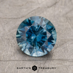 A 1.79-Carat Teal Blue Montana Sapphire (Heated) is displayed on a textured light gray surface with "Earth's Treasury" and a hexagon logo beneath.