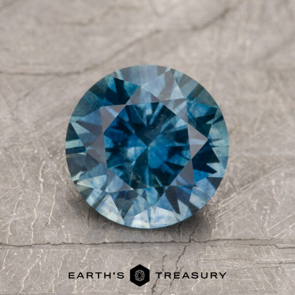 1.79-Carat Teal Blue Montana Sapphire (Heated) A 1.79-Carat Teal Blue Montana Sapphire (Heated) is displayed on a textured light gray surface with "Earth's Treasury" and a hexagon logo beneath.
