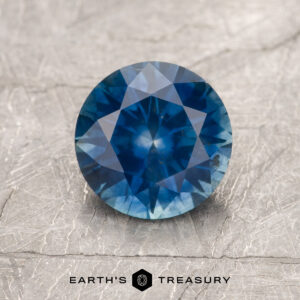A 1.64-carat rich blue Montana Sapphire (heated), brilliantly round-cut, glimmers on a textured gray surface. The "EARTH'S TREASURY" logo and text are displayed at the bottom of the image.