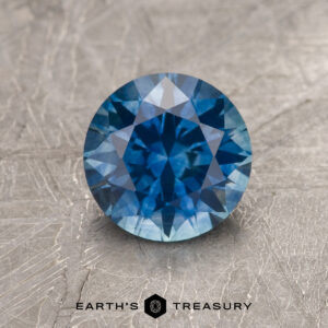 1.49-carat medium blue heated Montana Sapphire, round brilliant-cut, displayed on a textured background with the "Earth's Treasury" logo and text at the bottom.
