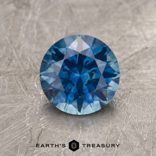 1.49-Carat Medium Blue Montana Sapphire (Heated) 1.49-carat medium blue heated Montana Sapphire, round brilliant-cut, displayed on a textured background with the "Earth's Treasury" logo and text at the bottom.