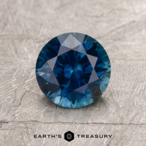 A 1.47-carat rich blue Montana Sapphire (heated), round and faceted, rests on a textured gray surface with “EARTH'S TREASURY” and a hexagonal logo shown below.