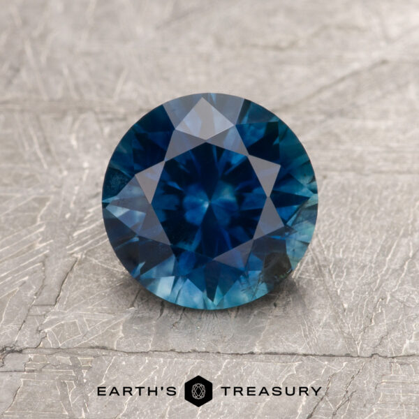 1.47-Carat Rich Blue Montana Sapphire (Heated) A 1.47-carat rich blue Montana Sapphire (heated), round and faceted, rests on a textured gray surface with “EARTH'S TREASURY” and a hexagonal logo shown below.