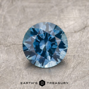 1.47-Carat Medium Blue Montana Sapphire (Heated), round brilliant-cut, showcased on a textured gray surface with the Earth's Treasury logo and text at the bottom.