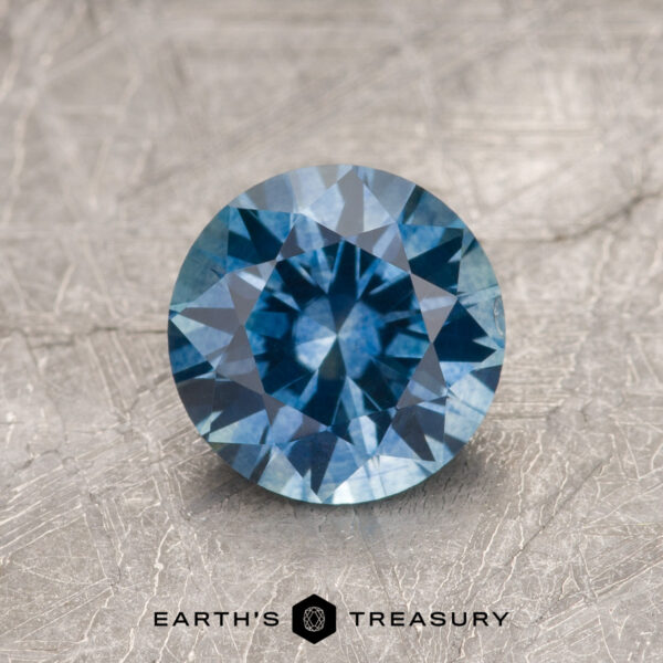 1.47-Carat Medium Blue Montana Sapphire (Heated) 1.47-Carat Medium Blue Montana Sapphire (Heated), round brilliant-cut, showcased on a textured gray surface with the Earth's Treasury logo and text at the bottom.