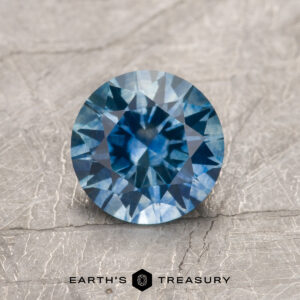 1.32-carat medium blue heated Montana sapphire with intricate facets shown on a textured gray surface; "Earth's Treasury" logo and text visible at the bottom.