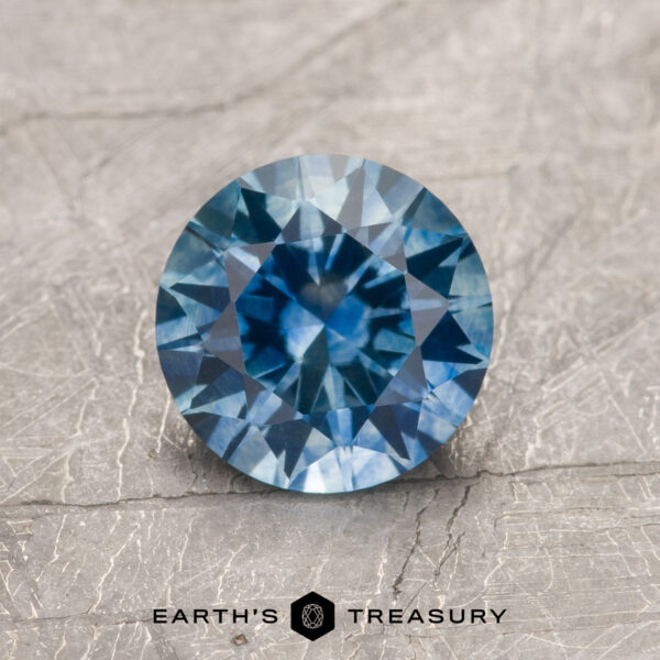 1.32-Carat Medium Blue Montana Sapphire (Heated) 1.32-carat medium blue heated Montana sapphire with intricate facets shown on a textured gray surface; "Earth's Treasury" logo and text visible at the bottom.