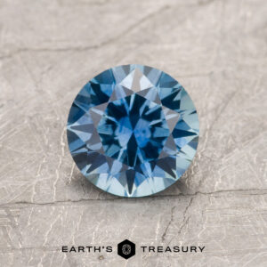 A 1.31-carat teal blue Montana Sapphire (heated) with a round, faceted cut rests on a textured light gray surface, with "EARTH'S TREASURY" written below.