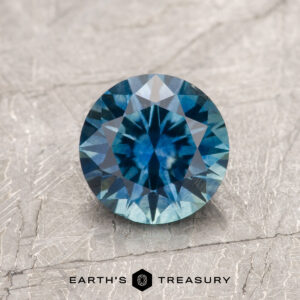 A 1.31-carat Teal Blue Montana Sapphire (Heated) with a round, faceted cut is shown on a textured light gray surface, accompanied by the text "EARTH'S TREASURY" below.