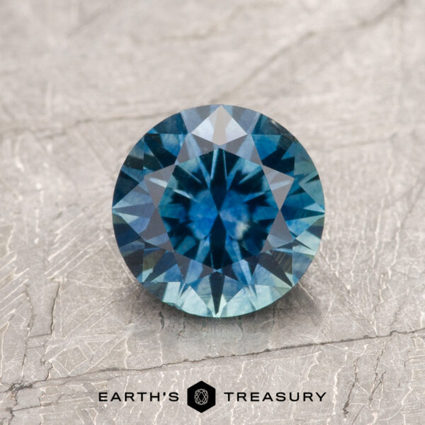 1.31-Carat Teal Blue Montana Sapphire (Heated) A 1.31-carat Teal Blue Montana Sapphire (Heated) with a round, faceted cut is shown on a textured light gray surface, accompanied by the text "EARTH'S TREASURY" below.
