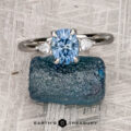 The "Merope" 3x2 ring in platinum, featuring a 1.08-carat Montana sapphire and two smaller stones, is displayed on a textured blue mineral with the words "Earth's Treasury" below.