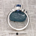 A platinum "Merope" ring featuring a 1.42-carat Ceylon sapphire and two clear side stones stands upright on a textured gray surface, with the "Earth's Treasury" logo below.
