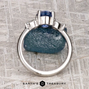 A platinum "Merope" ring featuring a 1.42-carat Ceylon sapphire and two clear side stones stands upright on a textured gray surface, with the "Earth's Treasury" logo below.