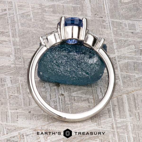 The "Merope" ring in platinum with 1.42-carat Ceylon sapphire A platinum "Merope" ring featuring a 1.42-carat Ceylon sapphire and two clear side stones stands upright on a textured gray surface, with the "Earth's Treasury" logo below.