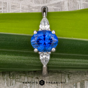 A platinum "Merope" ring featuring a 1.42-carat Ceylon sapphire and two pear-shaped clear stones, displayed on a green leaf background with "Earth's Treasury" branding below.