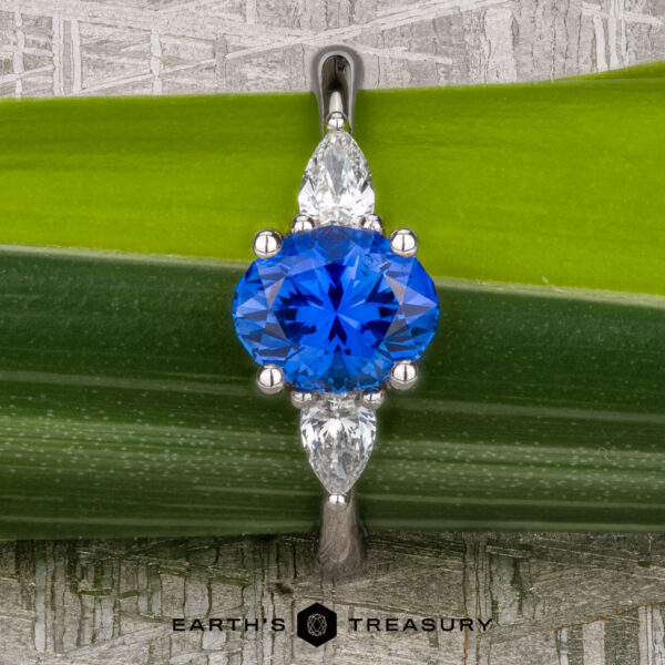 The "Merope" ring in platinum with 1.42-carat Ceylon sapphire A platinum "Merope" ring featuring a 1.42-carat Ceylon sapphire and two pear-shaped clear stones, displayed on a green leaf background with "Earth's Treasury" branding below.