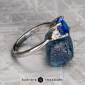 The "Merope" ring in platinum, featuring a 1.42-carat Ceylon sapphire center, is showcased on a rough blue gem against a geometric patterned backdrop.