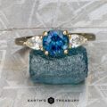The "Merope" 5x3 ring in 18K yellow gold, set with a 1.19-carat Montana sapphire and two pear-shaped white stones, rests on rough blue crystal against a textured background.