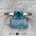 The "Clio" ring 4x2 in 14K white gold, featuring a 1.69-carat Montana sapphire and two baguette side stones, is displayed upright on rough blue mineral with the "Earth's Treasury" logo below.