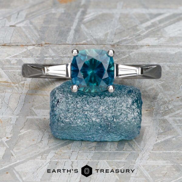 The "Clio" ring 4x2 in 14K white gold with 1.69-carat Montana sapphire The "Clio" ring 4x2 in 14K white gold, featuring a 1.69-carat Montana sapphire and two baguette side stones, is displayed upright on rough blue mineral with the "Earth's Treasury" logo below.