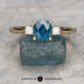 The "Wisteria" 2.5 ring in 14K yellow gold features a 1.18-carat oval Montana sapphire flanked by two round white stones, displayed on rough blue mineral with the "Earth's Treasury" logo below.