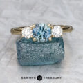 The "Wisteria" 3.5 ring in 14K yellow gold, featuring a 1.00-carat Montana sapphire and two white side stones, rests atop a rough blue crystal with the "Earth's Treasury" logo displayed below.