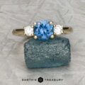 The "Wisteria" 3.0 ring in 14k yellow gold, featuring a 1.14-carat Montana sapphire and two white side stones, is displayed on a rough blue stone atop a textured gray surface. The “Earth’s Treasury” logo appears at the bottom.