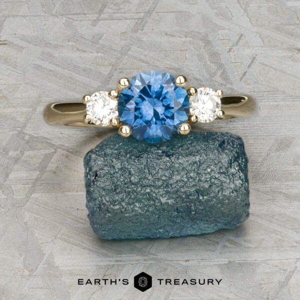 The "Wisteria" 3.0 ring in 14k yellow gold with 1.14-carat Montana sapphire The "Wisteria" 3.0 ring in 14k yellow gold, featuring a 1.14-carat Montana sapphire and two white side stones, is displayed on a rough blue stone atop a textured gray surface. The “Earth’s Treasury” logo appears at the bottom.
