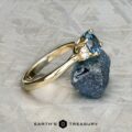 The "Wisteria" 3.0 ring in 14k yellow gold, featuring a 1.14-carat Montana sapphire and accent stones, is displayed beside a rough blue mineral on a patterned surface, with the Earth's Treasury logo at the bottom.