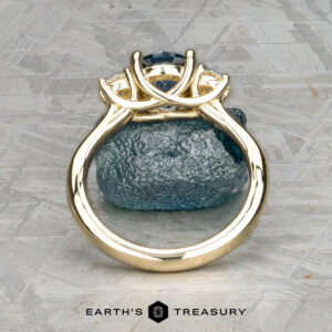 The "Wisteria" 3.0 ring in 14k yellow gold featuring a 1.14-carat Montana sapphire stands upright on a rough stone surface, viewed from the base, with the Earth's Treasury logo at the bottom.