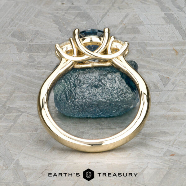 The "Wisteria" 3.0 ring in 14k yellow gold with 1.14-carat Montana sapphire The "Wisteria" 3.0 ring in 14k yellow gold featuring a 1.14-carat Montana sapphire stands upright on a rough stone surface, viewed from the base, with the Earth's Treasury logo at the bottom.