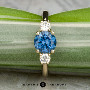 The "Wisteria" 3.0 ring in 14k yellow gold features a 1.14-carat Montana sapphire between two white diamonds, shown against green leaves with the "Earth's Treasury" logo at the bottom.