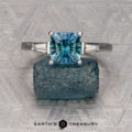 The Le Renard, Yann three-stone ring with a blue sapphire and baguette side stones is displayed on a rough blue gem over a patterned gray background featuring the Earth’s Treasury logo.