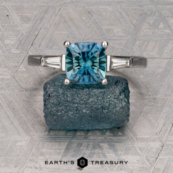 Le Renard, Yann The Le Renard, Yann three-stone ring with a blue sapphire and baguette side stones is displayed on a rough blue gem over a patterned gray background featuring the Earth’s Treasury logo.