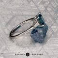 A Le Renard, Yann three-stone ring with a blue gemstone and two clear side stones is displayed on a rough blue mineral, set against a geometric patterned background. The "Earth's Treasury" logo appears below.