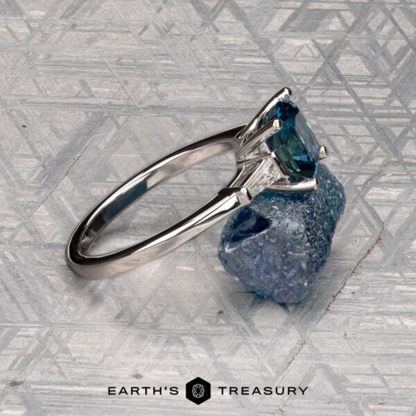 Le Renard, Yann A Le Renard, Yann three-stone ring with a blue gemstone and two clear side stones is displayed on a rough blue mineral, set against a geometric patterned background. The "Earth's Treasury" logo appears below.