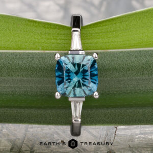 A Le Renard, Yann engagement ring with a blue gemstone cushion cut center and baguette side stones is displayed on a green leaf; the "Earth's Treasury" logo appears at the bottom.