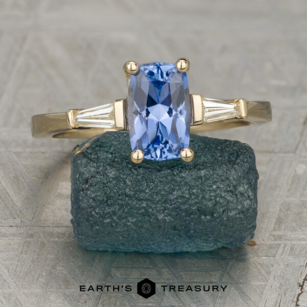 The "Clio" ring in 14K yellow gold with 1.09-carat Ceylon sapphire The "Clio" ring in 14K yellow gold, featuring a 1.09-carat Ceylon sapphire and two tapered side stones, stands upright on rough blue crystal with the Earth's Treasury logo beneath.