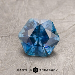A 1.33-carat medium blue heated Montana Sapphire with a hexagonal cut rests on a textured gray surface. "Earth's Treasury" and the logo are shown at the image’s bottom.