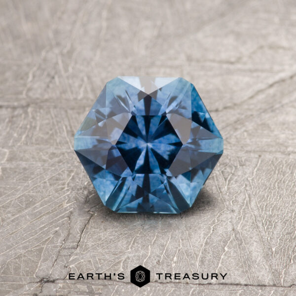 1.27-Carat Medium Blue Montana Sapphire (Heated) A 1.27-carat medium blue Montana sapphire (heated) with intricate facets is displayed on a textured gray surface, with the Earth's Treasury logo beneath it.