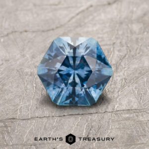 A 1.25-carat teal blue Montana sapphire (heated), cut in an octagonal shape, rests on a textured gray surface with the "Earth's Treasury" logo beneath.
