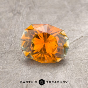 A 1.60-carat orange-yellow particolored Montana sapphire (heated) is displayed on a textured gray surface featuring the Earth's Treasury logo at the bottom.