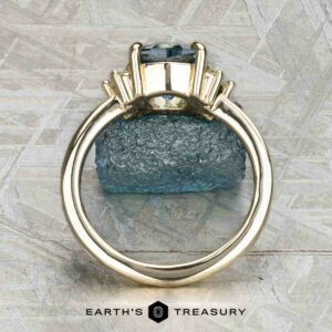 The "Hesperia" ring in 14K yellow gold showcases a 2.21-carat Montana sapphire and side baguette stones, shown upright with a rough blue mineral and the Earth’s Treasury logo in the background.