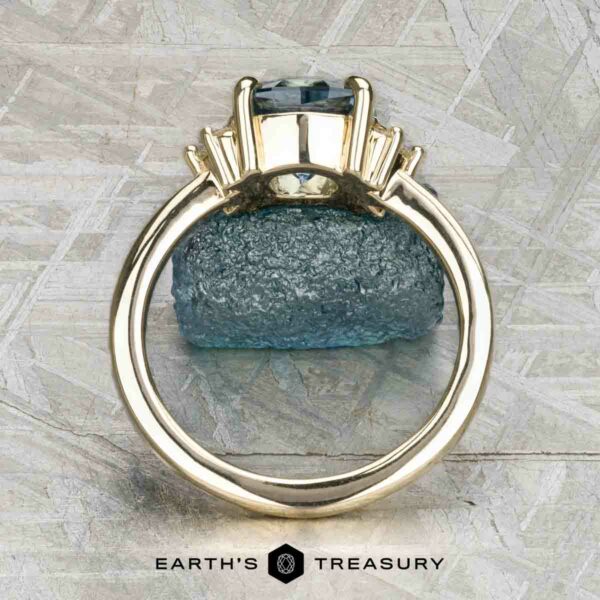 The "Hesperia" ring in 14K yellow gold with 2.21-carat Montana sapphire The "Hesperia" ring in 14K yellow gold showcases a 2.21-carat Montana sapphire and side baguette stones, shown upright with a rough blue mineral and the Earth’s Treasury logo in the background.