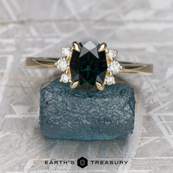 The "Hesperia" ring in 14K yellow gold with 1.55-carat Australian sapphire The "Hesperia" ring in 14K yellow gold showcases a 1.55-carat Australian sapphire with accent diamonds, displayed on a rough bluish stone above the "EARTH'S TREASURY" logo.