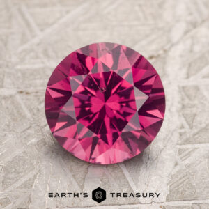 A 2.26-carat magenta pink Burmese spinel with intricate faceting is displayed on a textured gray surface, featuring the logo "Earth's Treasury" beneath it.