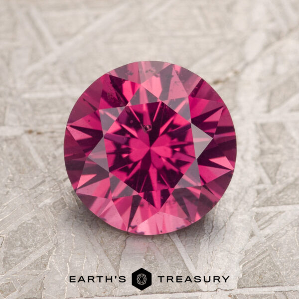A 2.26-carat magenta pink Burmese spinel with intricate faceting is displayed on a textured gray surface, featuring the logo "Earth's Treasury" beneath it.