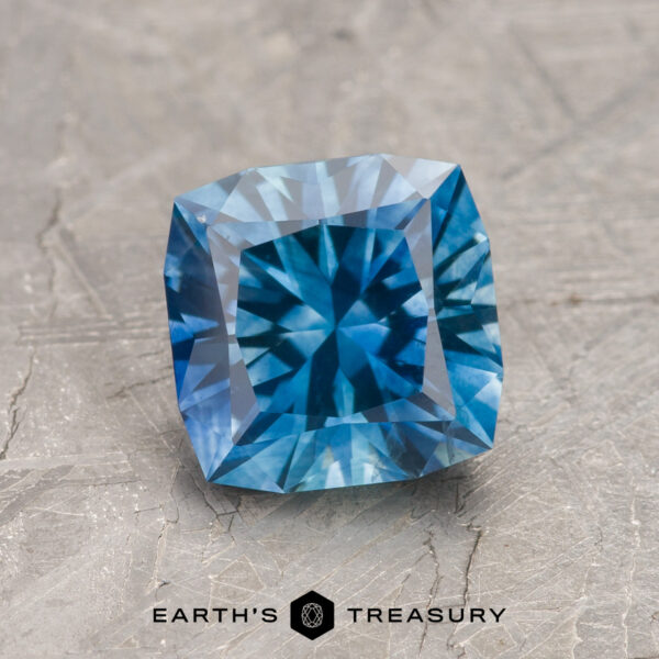1.89-Carat Medium Blue Montana Sapphire (Heated) A 1.89-carat medium blue heated Montana Sapphire with intricate facets is displayed on a textured surface, with the "EARTH'S TREASURY" logo visible below the gem.